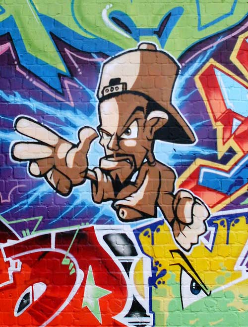 Graffito auf einer Backsteinwand. Das Bild zeigt einen comichaft stilisierten, posierenden Rapper mit umgedrehter Baseballmütze.