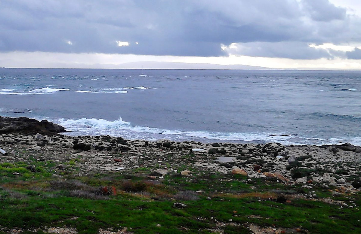 Blick an der Küste von Tarifa nach Marokko. Der Himmel ist bedeckt, das Wasser dunkel. In der Ferne: Die Umrisse von Bergen, die aus dem Wasser ragen.