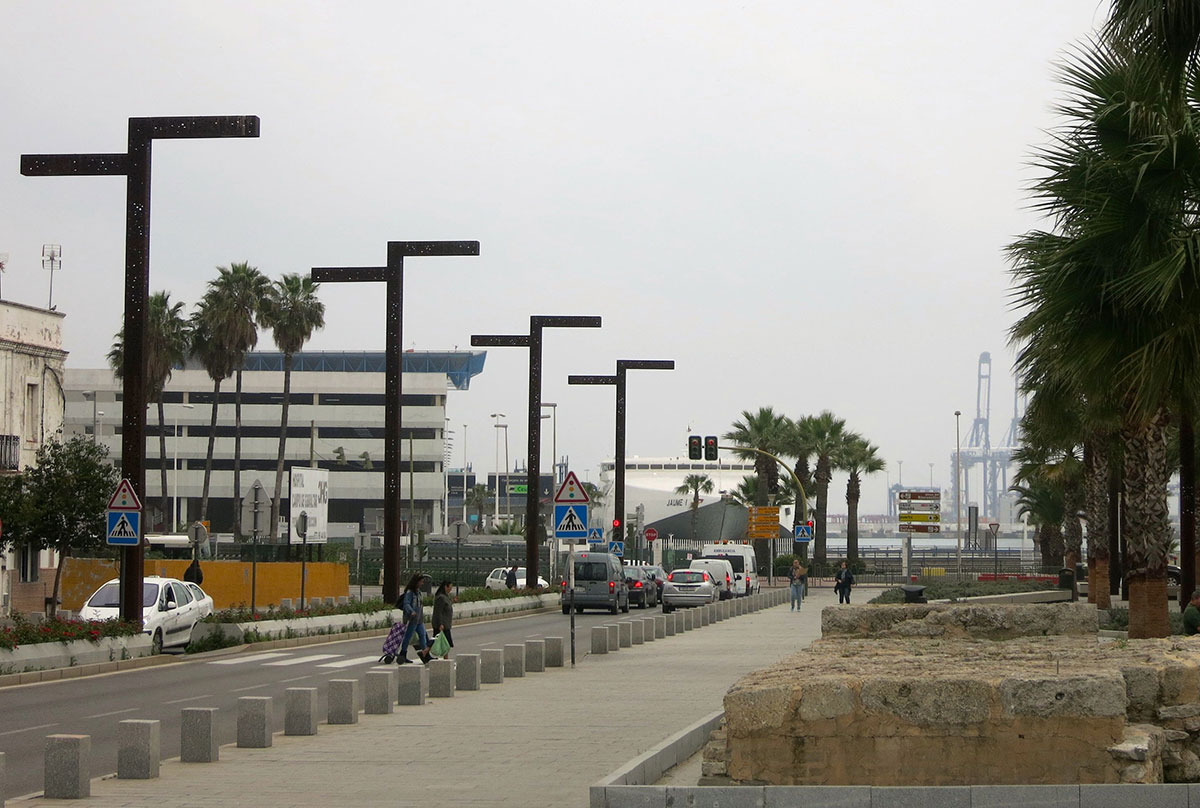 Strasse in der Stadt Algeciras. Lampen und Palmen säumen die fast menschenleere Allee. Im Hintergrund: Autos, ein ankerndes Schiff, sowie Kräne und Gebäude des Hafens.