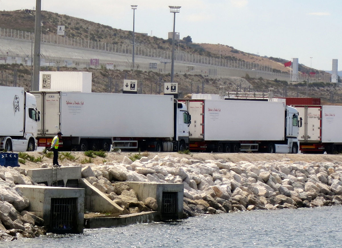 Im Hafen von Tanger, Marrokko: Eine Kolonne von Lastwagen wartet auf einer Strasse direkt am Meer darauf, verschifft zu werden. Ein Mann in einer grünen Weste steht daneben und schaut ins Meer. Im Hintergrund: Kahle Hügel und Grenzanlagen (Wachtürme, Zäune).
