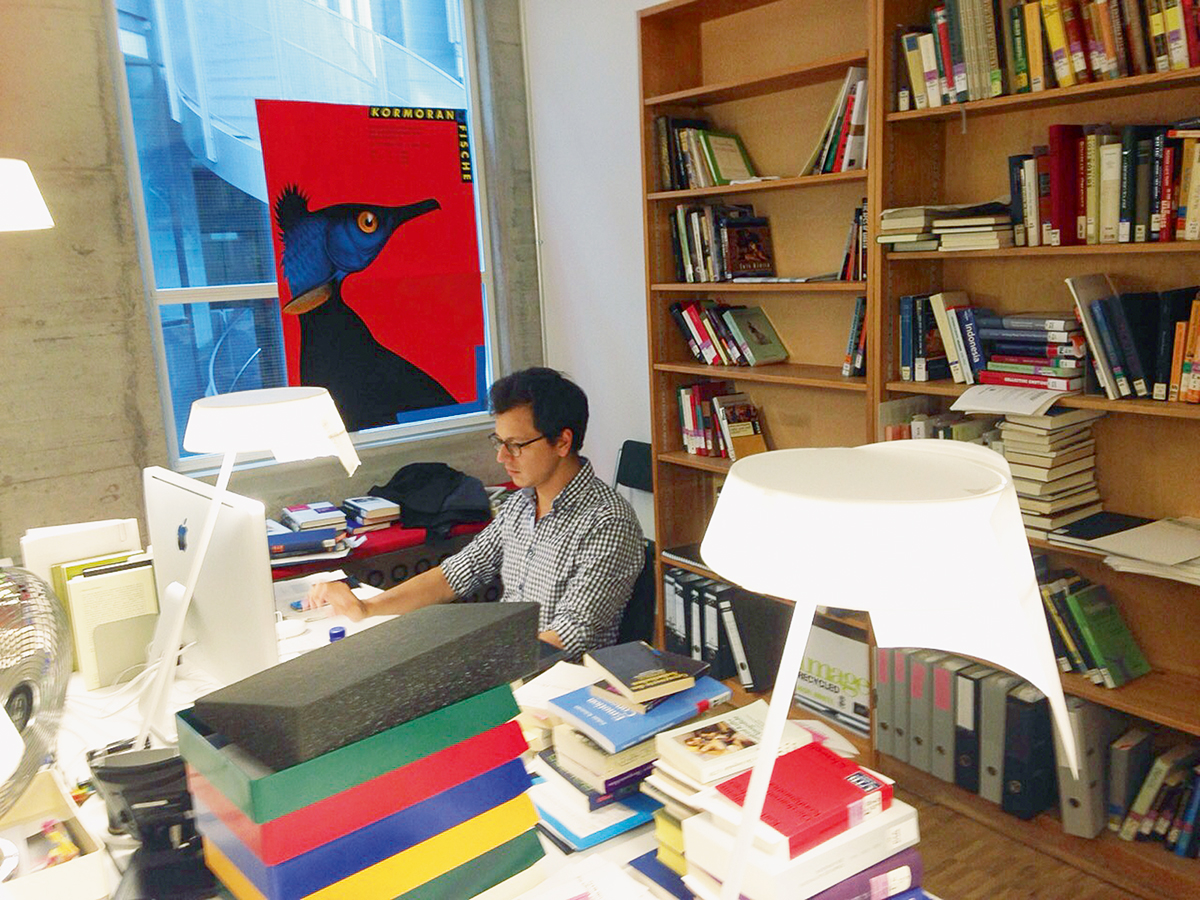 Fermin Suter sitzt am Schreibtisch in seinem Büro in der Unitobler und arbeitet am Computer. Auf dem Tisch und in den Regalen stapeln sich Bücher und Ordner. Am Fenster im Hintergrund hängt das Poster eines Vogels.