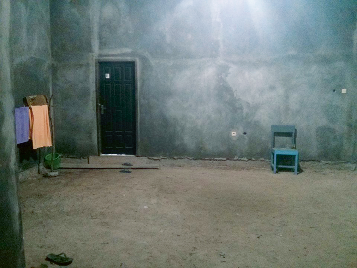 Unterkunft in Indonesien: Ein kahler Raum mit unverputzten Wänden. An der Wand, neben der Tür, steht ein kleiner Stuhl. Auf einem Gestell hängen Tücher, daneben steht ein Eimer.