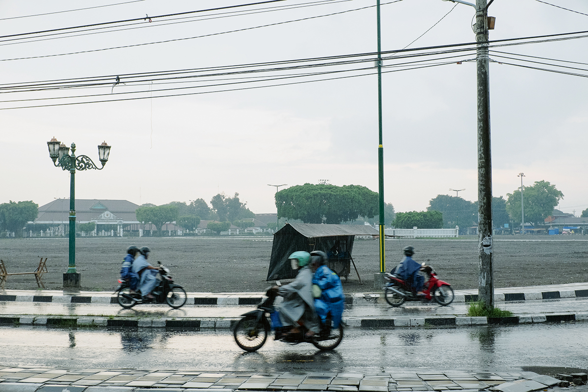 Strasse in Yogyakarta: Menschen auf Mofas fahren durch den strömenden Regen. Der Himmel ist grau und wird vom nassen Asphal gespiegelt. Elektorleitungen hängen über der Strasse. Im Hintergrund sind eine Wiese, Gebäude und Bäume zu sehen.