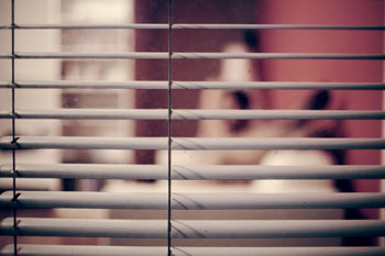 Blick durch Sonnenstoren in ein Zimmer, in dem eine Frau auf dem Bett sitzt (aus der Perspektive eines Stalkers)